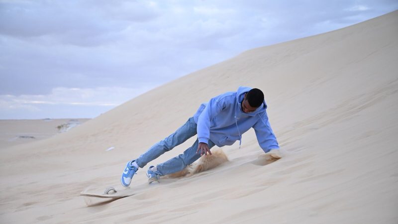 A young man in hoodie sandboarding down dunes in Egypt's desert landscape.