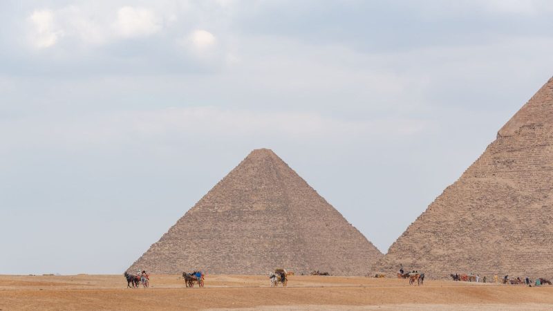 Camel riders near the iconic pyramids of Giza under a cloudy sky, showcasing Egypt's historical allure.