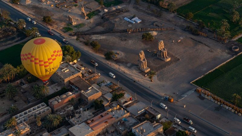 Hot air balloon flies over Egyptian landscape near ancient statues at sunset.