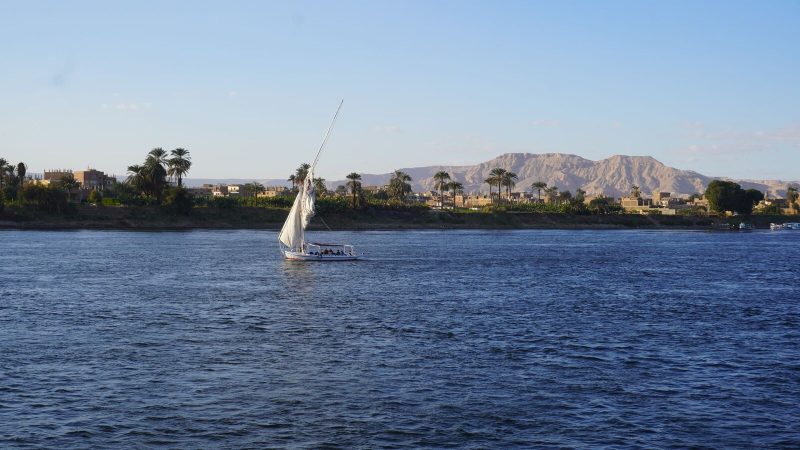 Scenic view of a traditional sailboat navigating the Nile at sunset, with lush riverbanks and distant mountains.