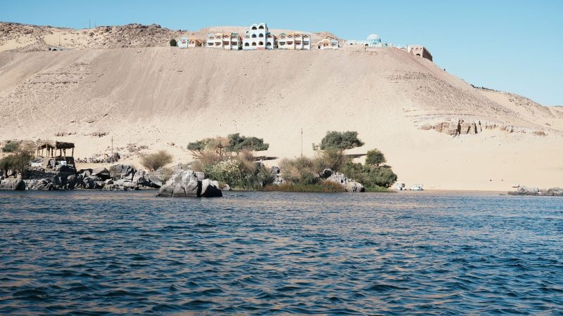 A tranquil desert scene featuring a hillside village overlooking a calm river under clear blue skies.