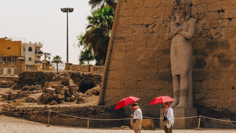 Two tourists walk past an ancient statue at Luxor Temple, Egypt, carrying red umbrellas.