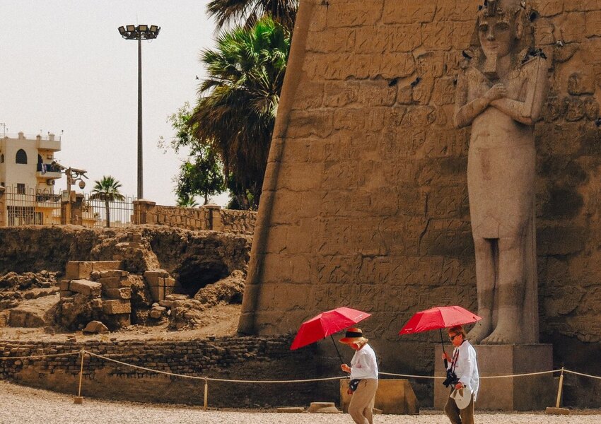 Two tourists walk past an ancient statue at Luxor Temple, Egypt, carrying red umbrellas.