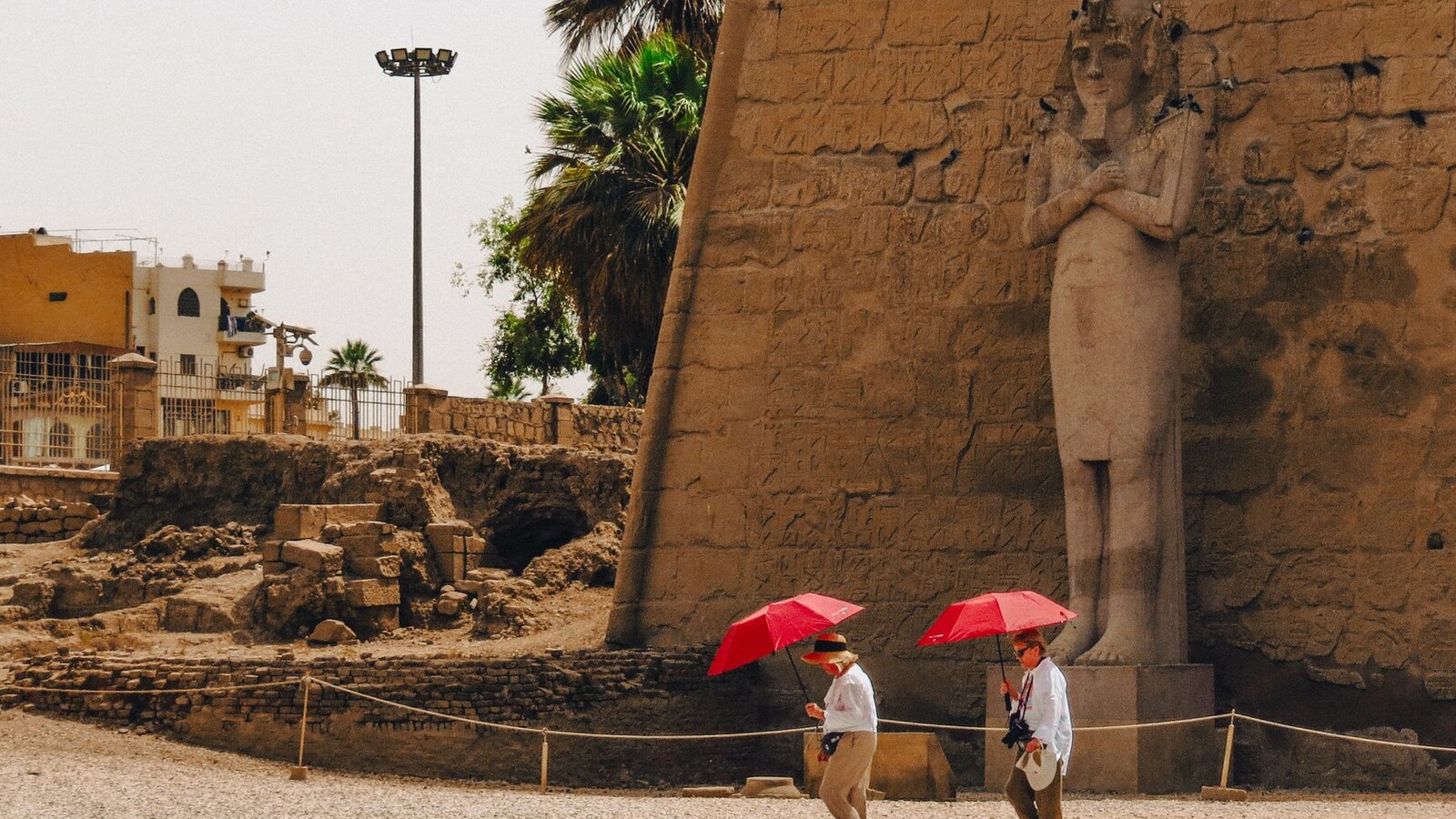 Two tourists walk past an ancient statue at Luxor Temple, Egypt, carrying red umbrellas.