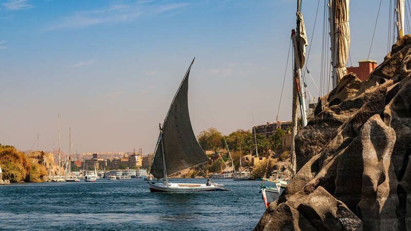 A traditional sailboat glides on the scenic Nile River near Aswan, Egypt, showcasing a tranquil landscape.