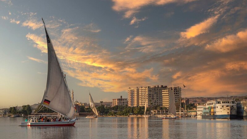 A traditional felucca sails the Nile at sunset with cityscape in the background.