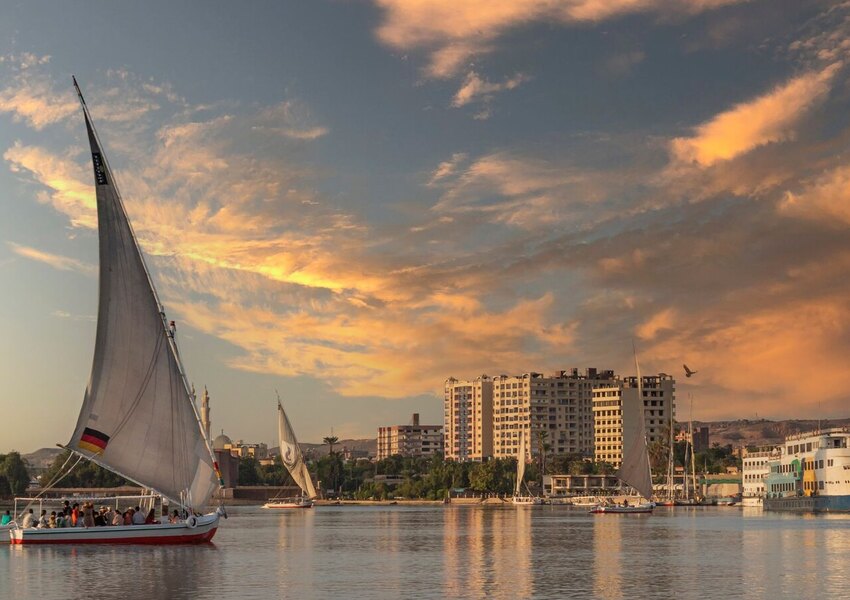 A traditional felucca sails the Nile at sunset with cityscape in the background.