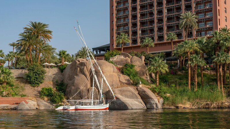 A serene sailboat rests on a calm river near boulders and lush palm trees, with a hotel in the background.